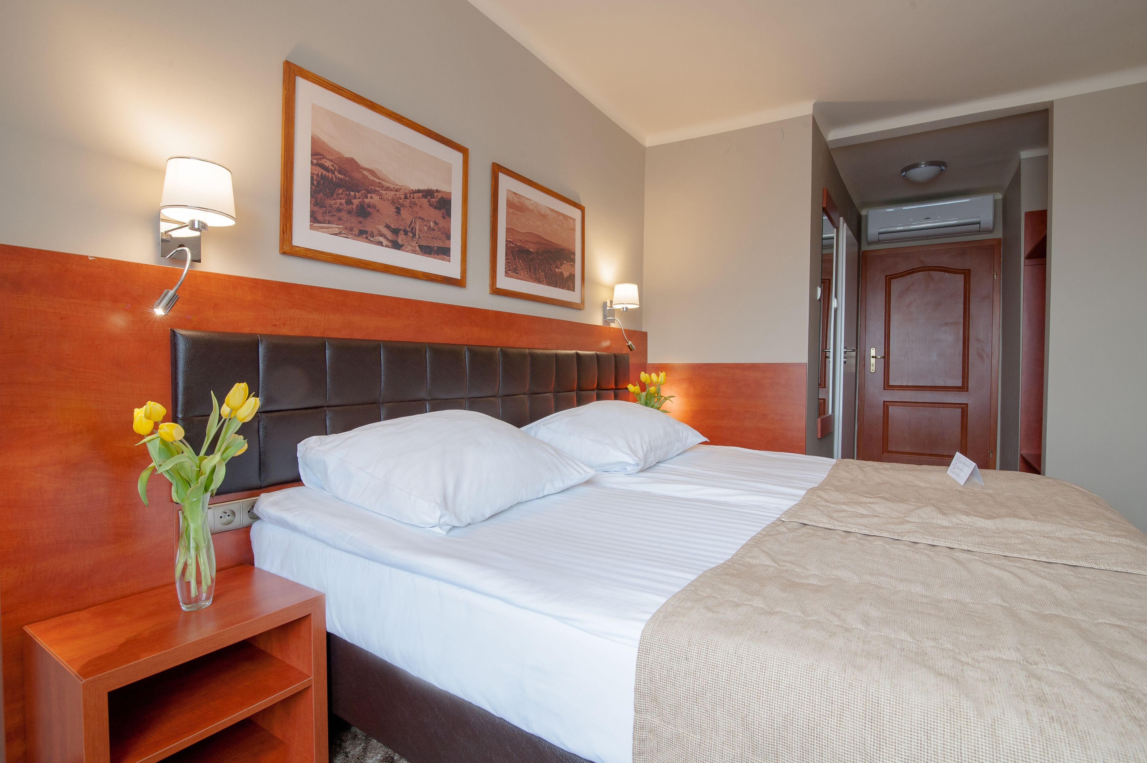 A hotel room with a large bed, a wooden headboard, two framed pictures on the wall, a nightstand with a vase of flowers, and a door leading to another room.