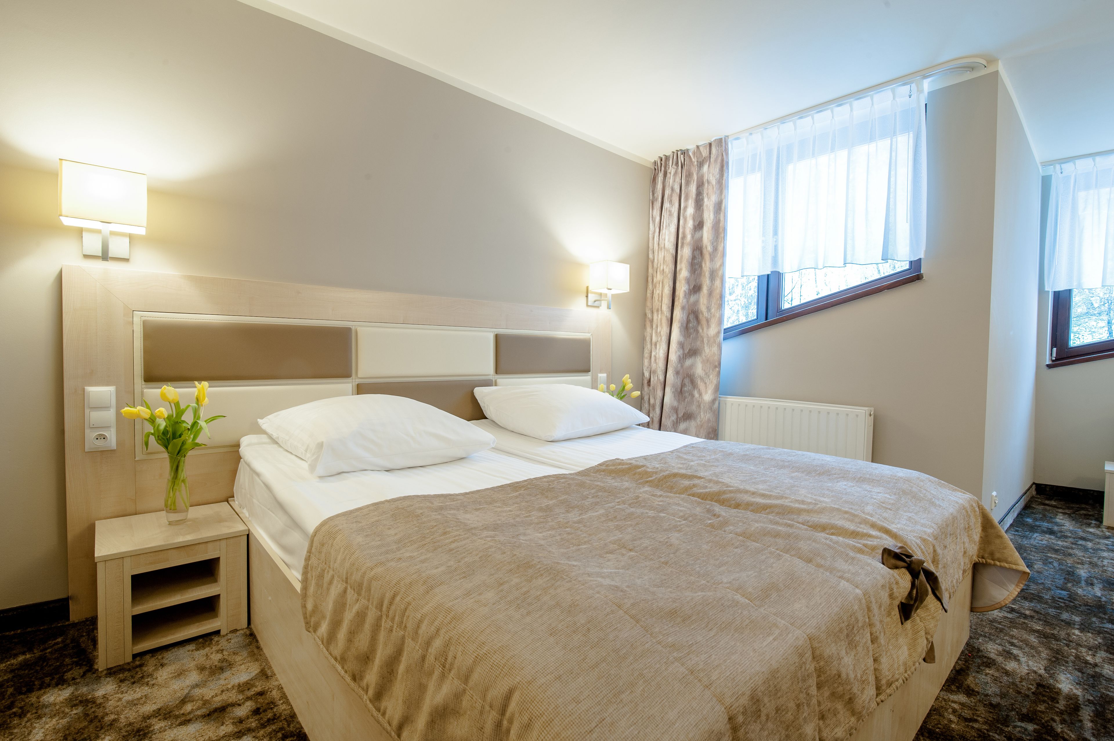 A hotel room with a large bed, bedside lamps, a nightstand, a window with curtains, and a radiator.