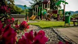Un terrain de jeux avec des toboggans et des structures d’escalade, entouré d’un jardin où poussent des fleurs roses.