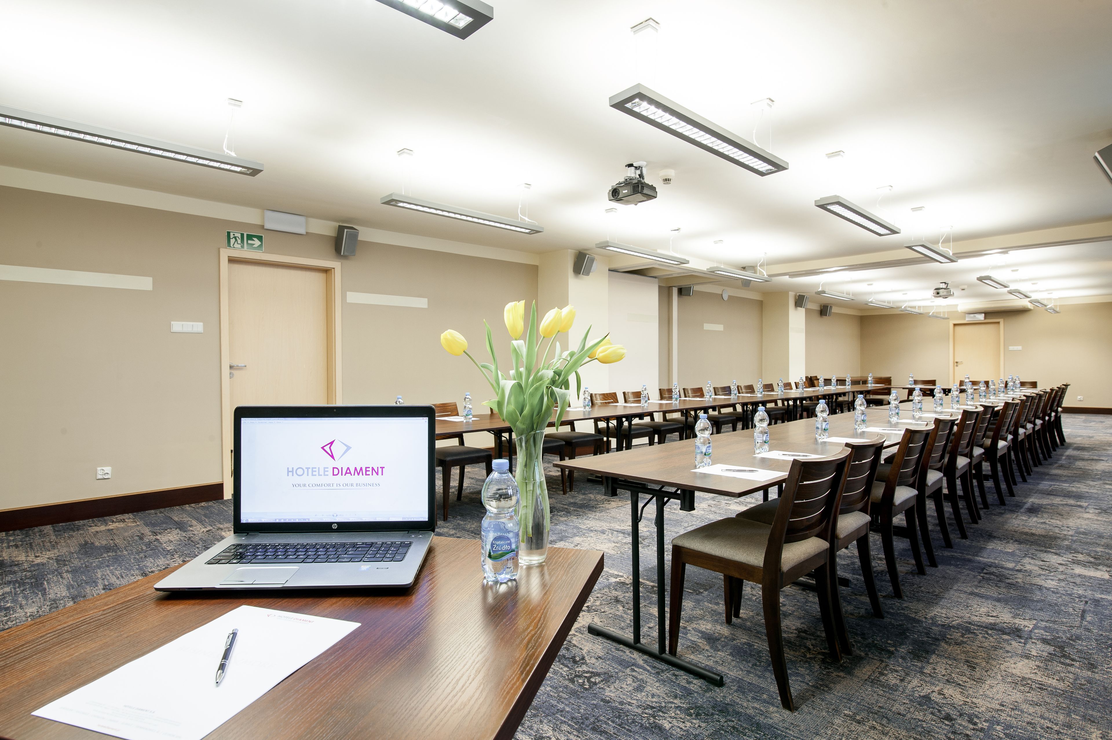 A conference room with a laptop displaying 'HOTEL DIAMOND' on the screen, a vase with yellow tulips, water bottles, and a carpeted floor.