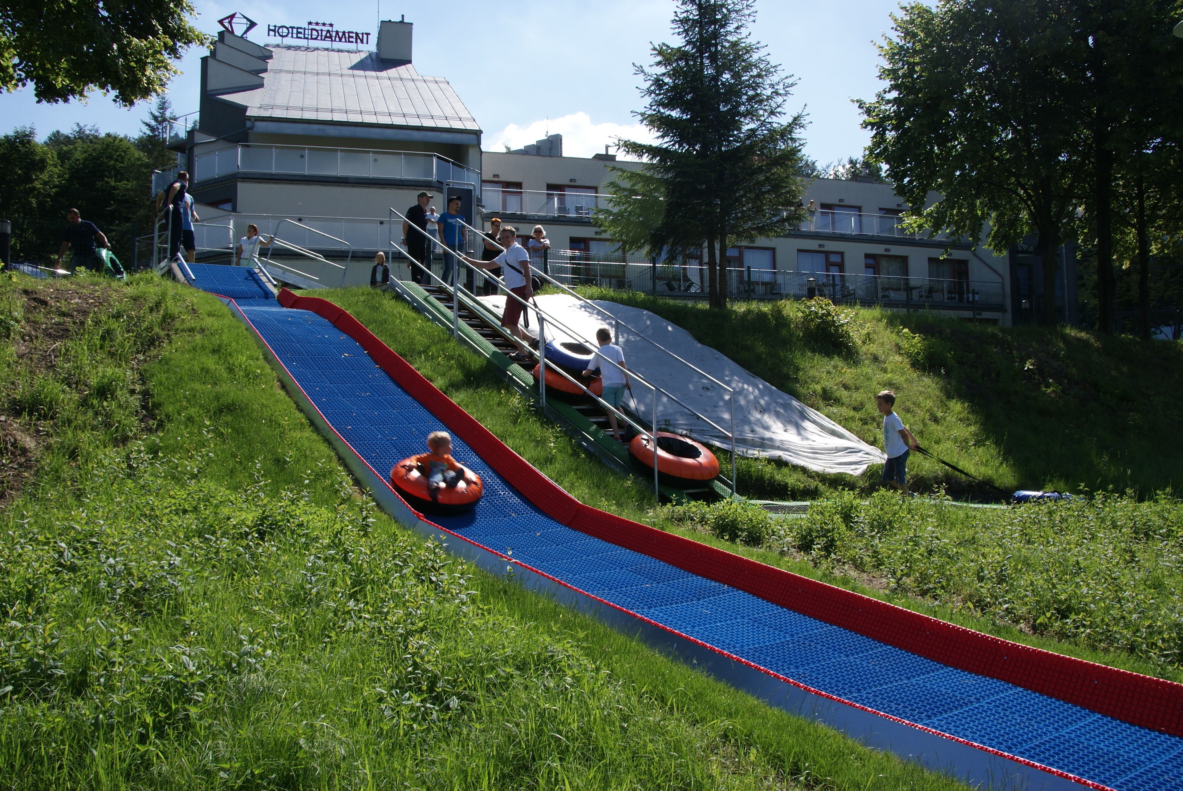 A water slide with inflatable tubes, surrounded by greenery and a building with a sign reading "HOTEL DIAMANT".