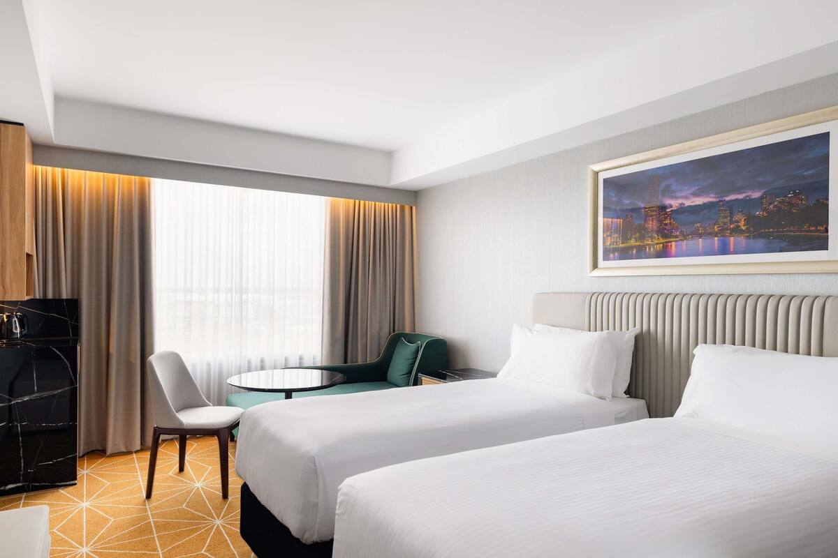 A hotel room with two beds, a desk, a chair, and a large window with curtains.