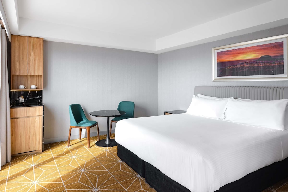 A hotel room with a bed, a small table, two chairs, a wooden cabinet, and a cityscape painting on the wall.