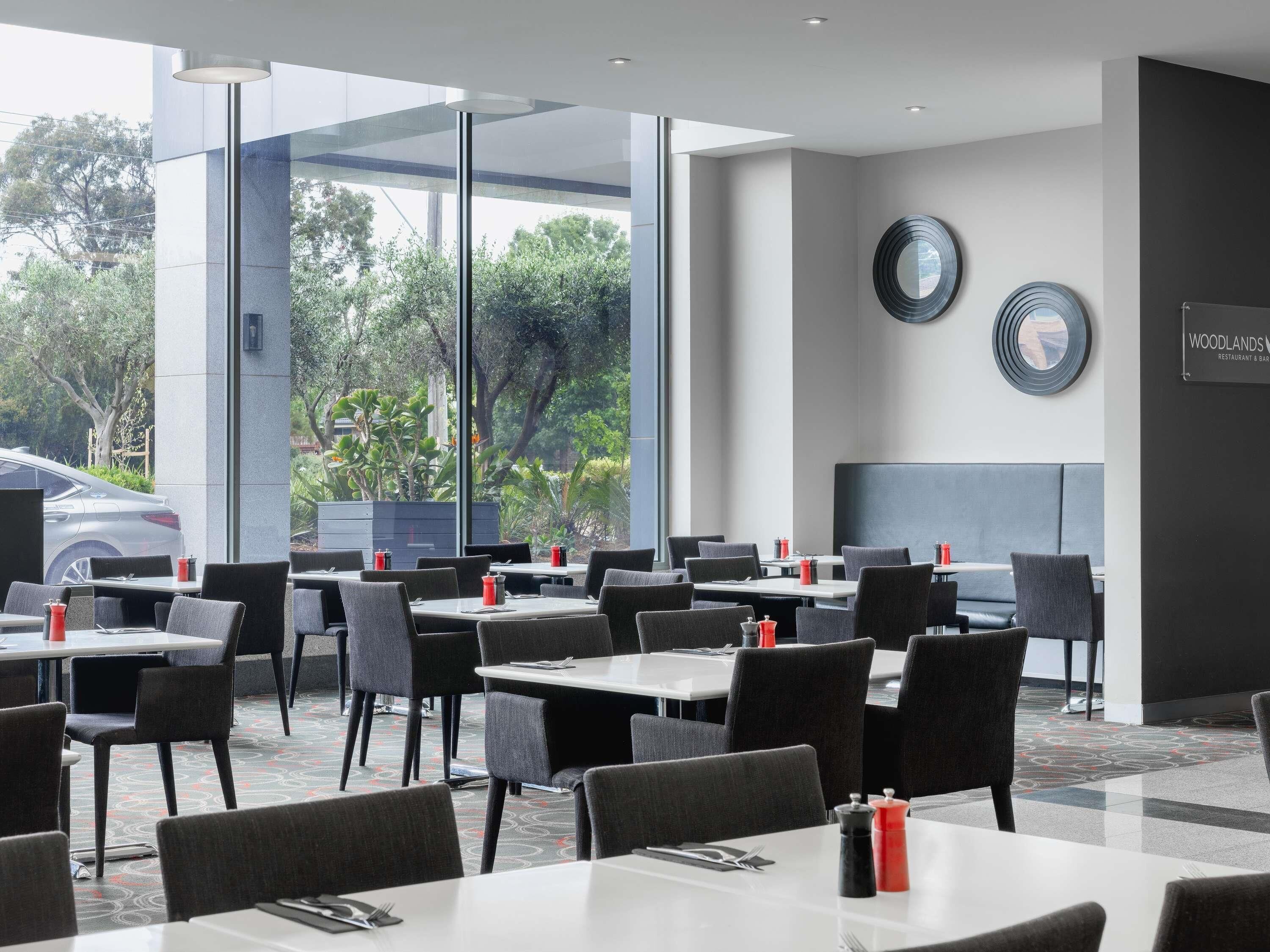 Een modern restaurant met tafels en stoelen, grote ramen en uitzicht op bomen en een auto.