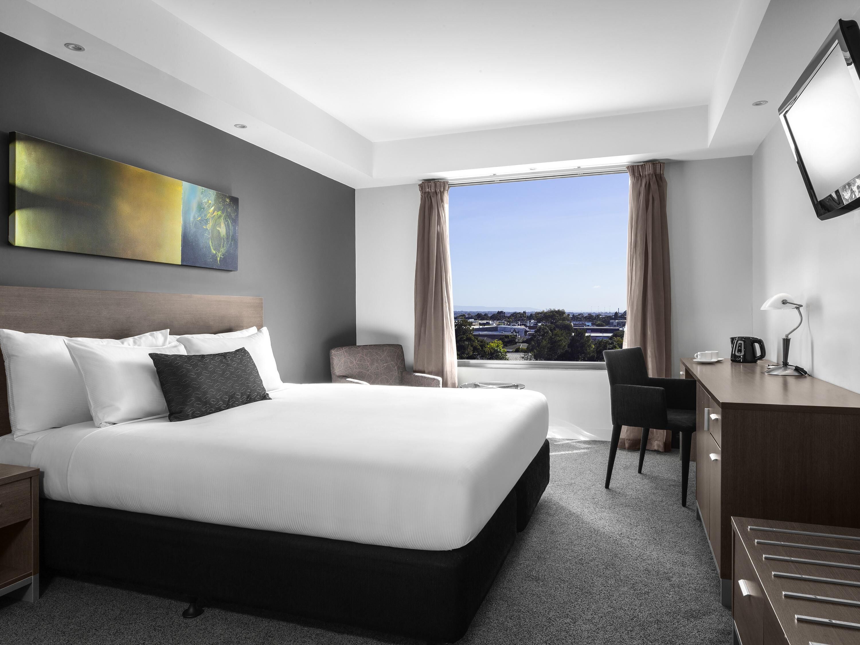 A hotel room with a large bed, a desk, a chair, a view of the city, and a painting on the wall.