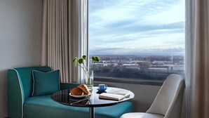 A modern hotel room with a teal sofa, a round table with a croissant, a cup, and a book, and a view of a cityscape through large windows. - Mantra Melbourne Airport (Tullamarine)