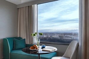 Habitación de hotel moderna con un sofá color turquesa, una mesa redonda con un croissant, una taza y un libro, y vistas de la ciudad a través de amplias ventanas.
