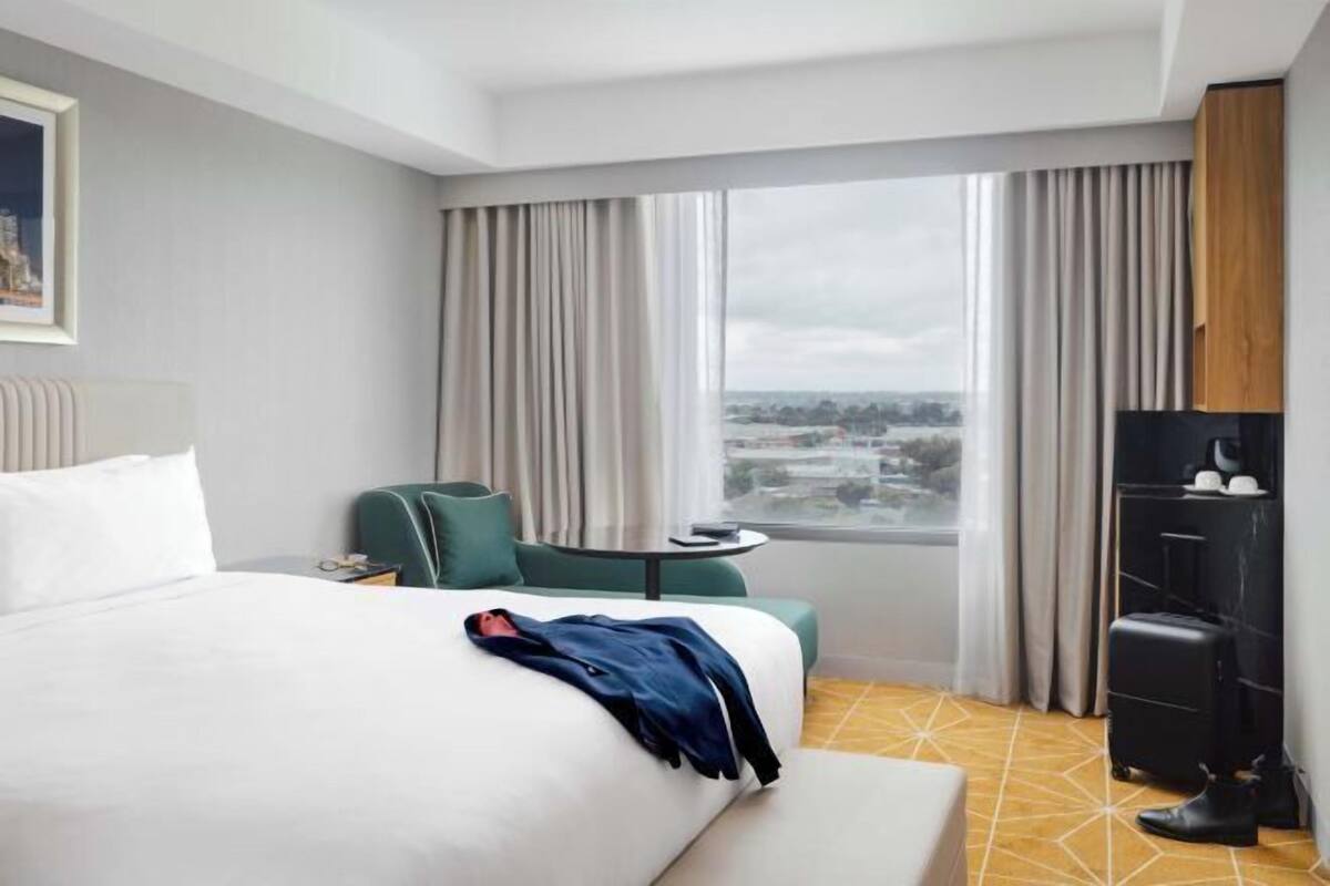 A hotel room with a bed, a small table, a chair, a wardrobe, and a window with a city view.