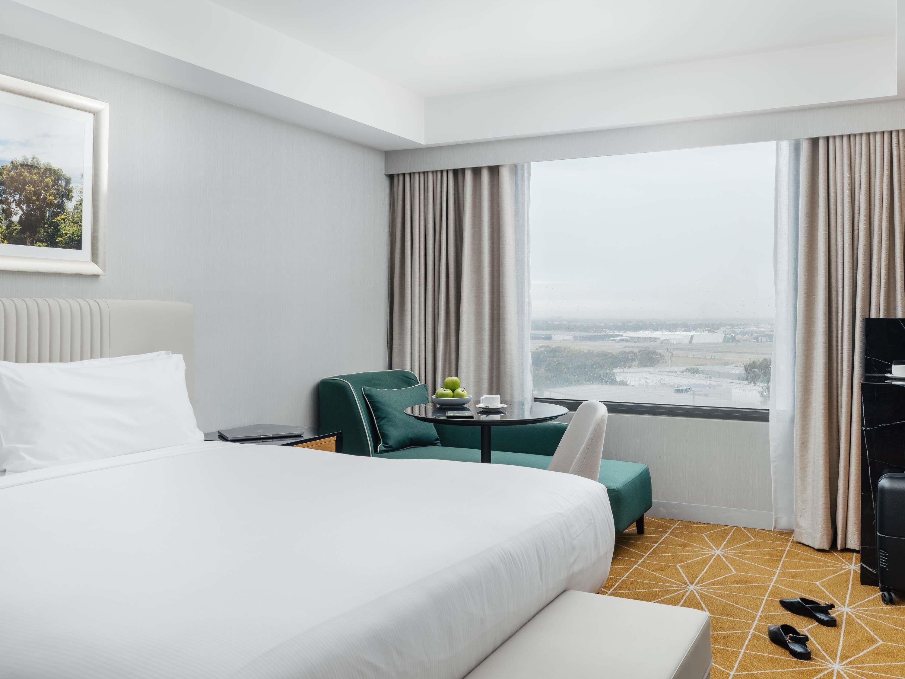 Een hotelkamer met een groot bed, een groene fauteuil, een klein tafeltje met een schaal fruit en uitzicht op de stad door grote ramen.