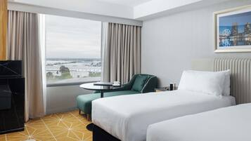 A hotel room with two beds, a desk, a chair, and a large window with a view of a cityscape.