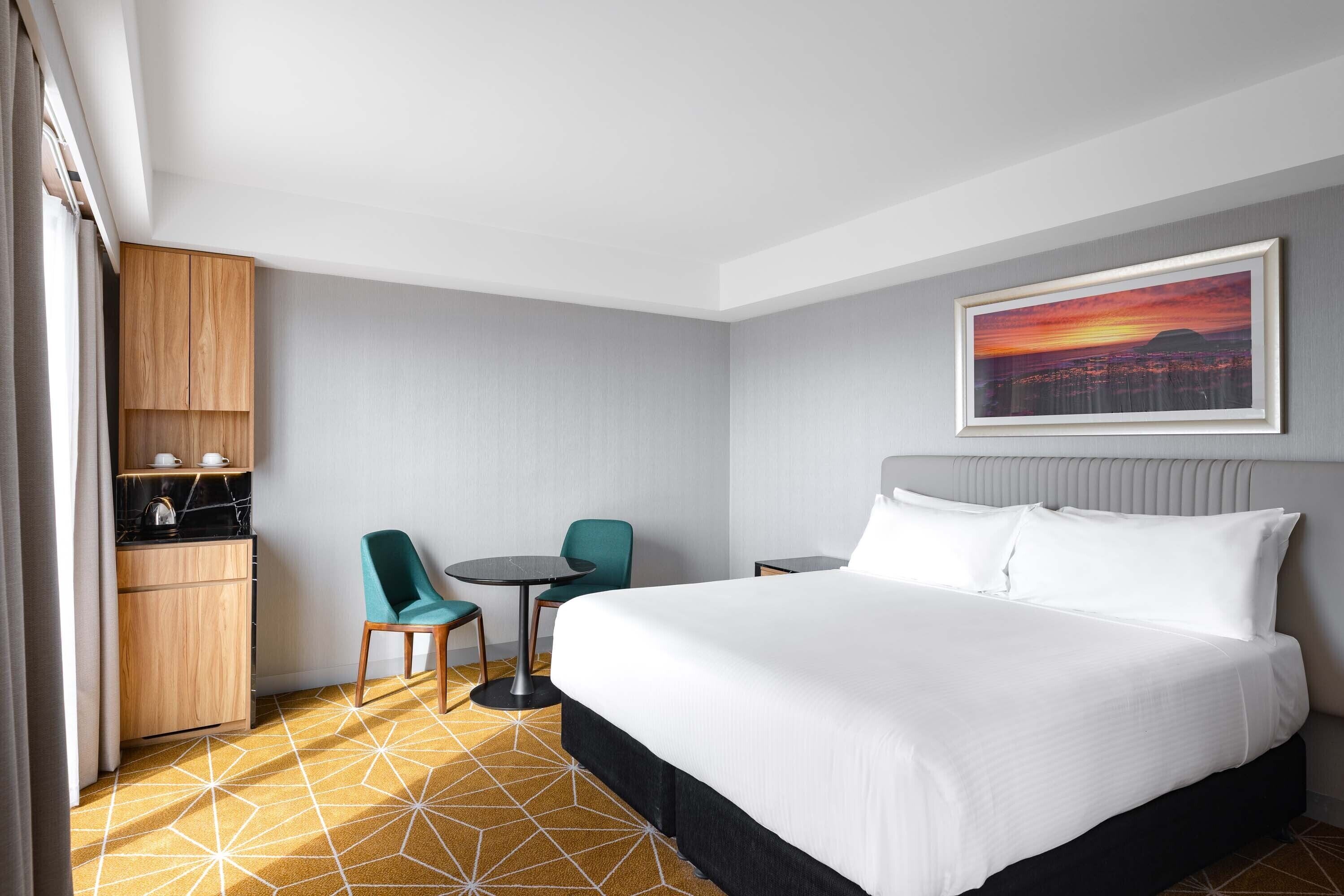 A hotel room with a large bed, a desk, a chair, a small table, and a kitchen area.
