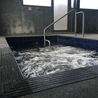 Un jacuzzi con una escalera de metal, rodeado de azulejos azules y una pared de hormigón.