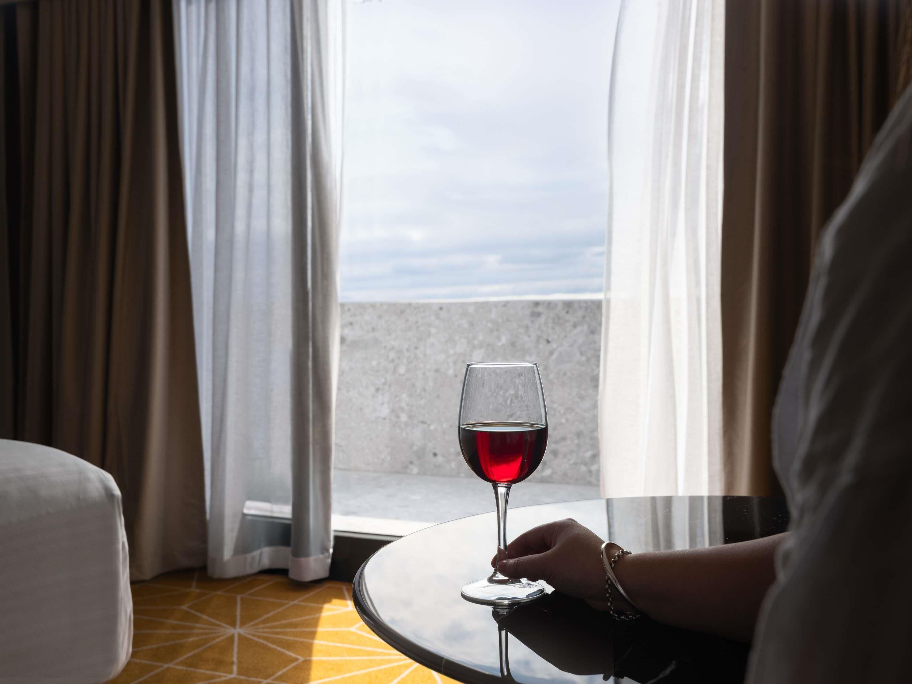 Una habitación con vistas a través de cortinas, una copa de vino tinto sobre una mesa y una persona sosteniendo la copa.