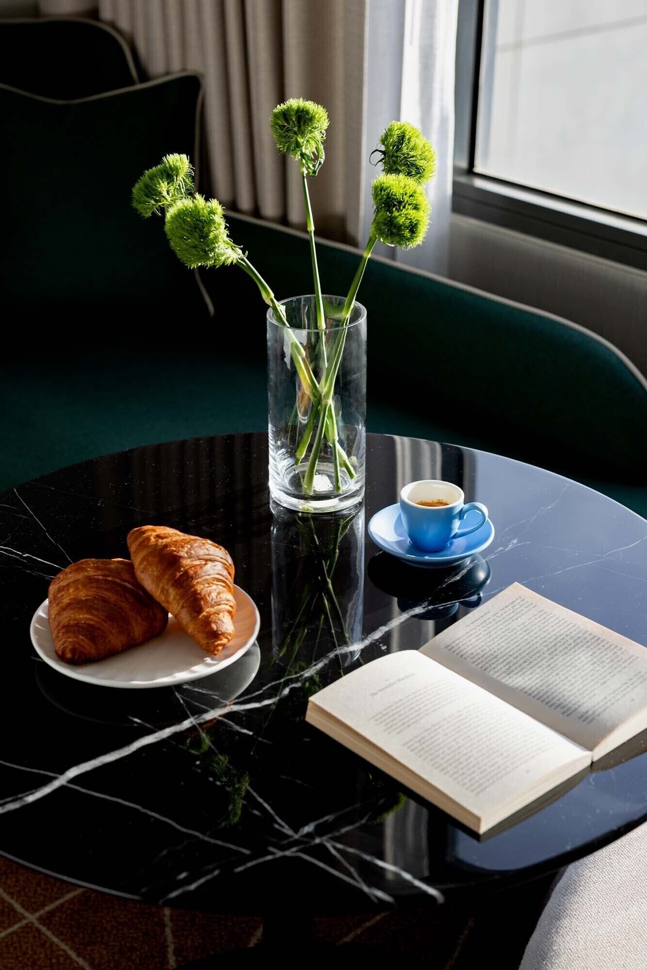 Une table avec des croissants, une tasse de café, un livre et un vase rempli de fleurs vertes.