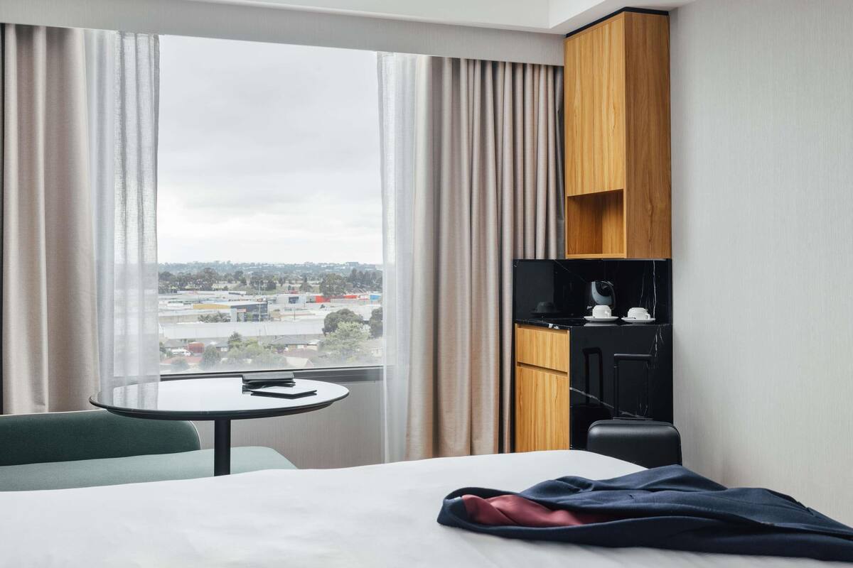 A hotel room with a bed, a small table, a chair, a wooden cabinet, and a view of a cityscape through large windows.