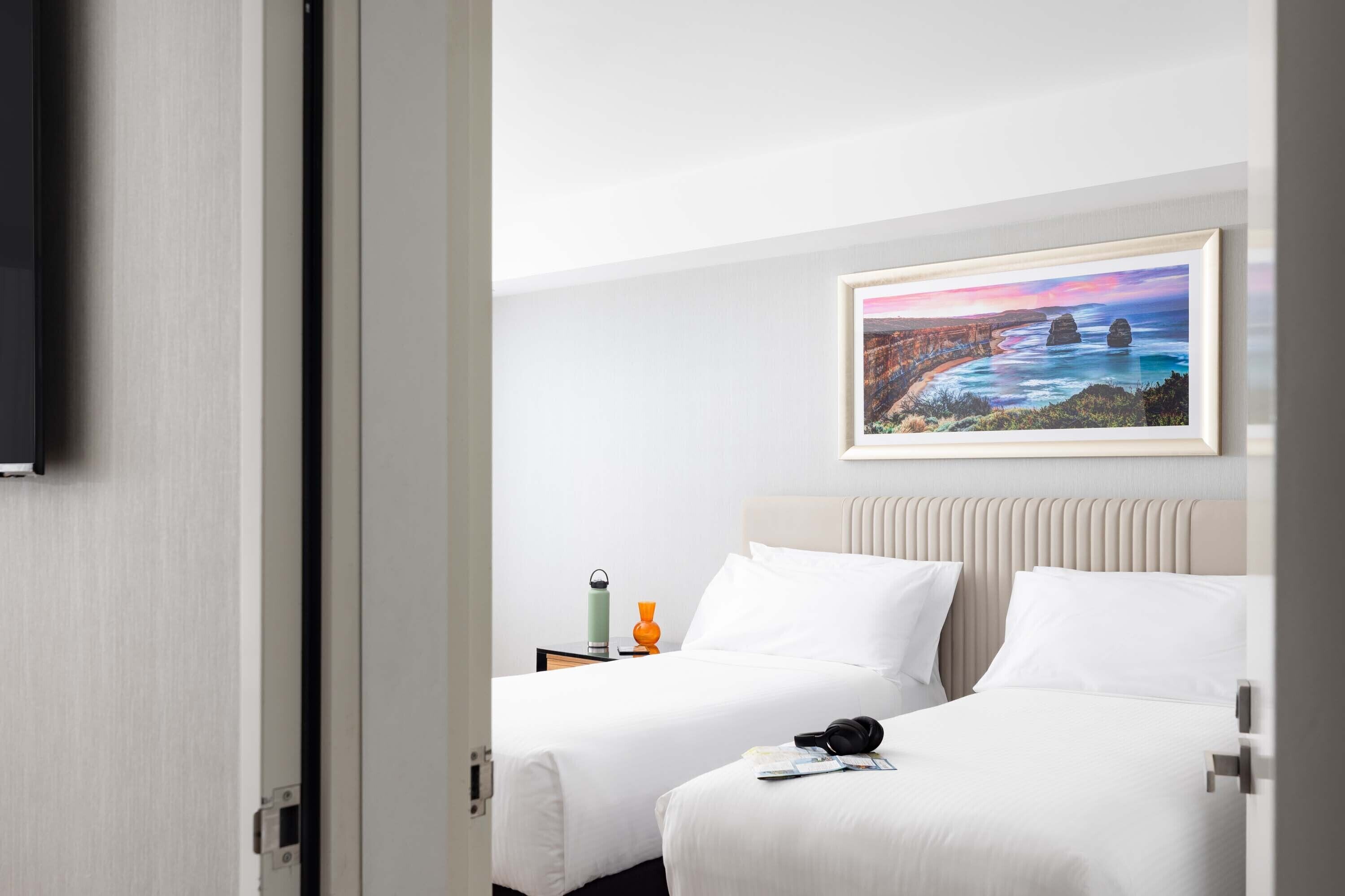 Een hotelkamer met twee bedden, een schilderij van een kustlandschap, een televisie aan de muur en een nachtkastje met een waterfles en een vaas.