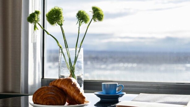 A table with a croissant, a blue cup, and a book, with a vase of green flowers and a glass of water on a table near a window with a view. - Mantra Melbourne Airport (Tullamarine)