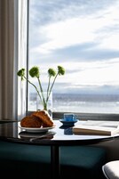 Una mesa con un croissant, una taza azul, un libro, un jarrón de flores verdes y un vaso de agua, cerca de una ventana con vistas.