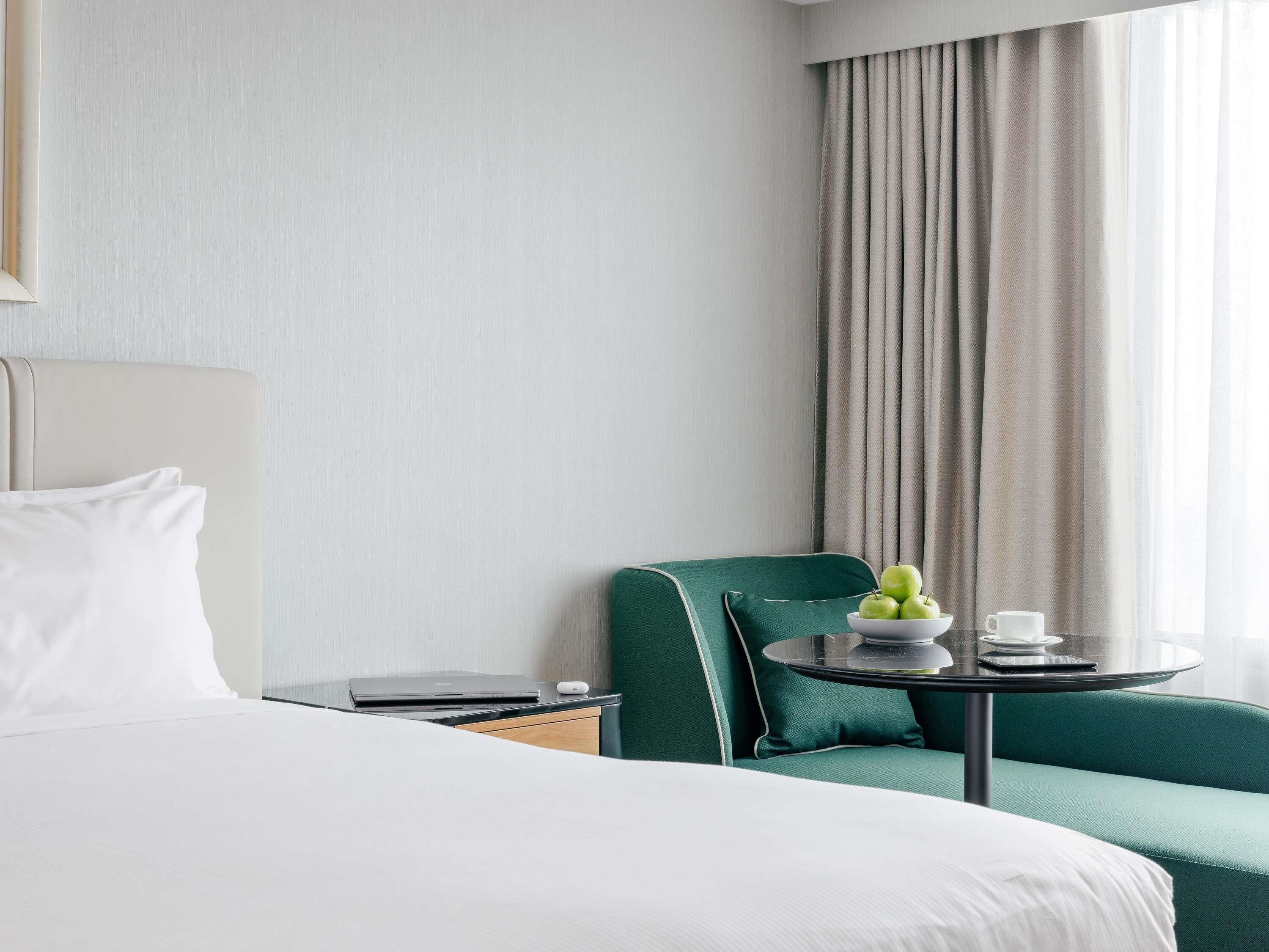 Una habitación de hotel con una cama, un sillón verde, una mesita con un frutero y un ordenador portátil sobre una mesita de noche.