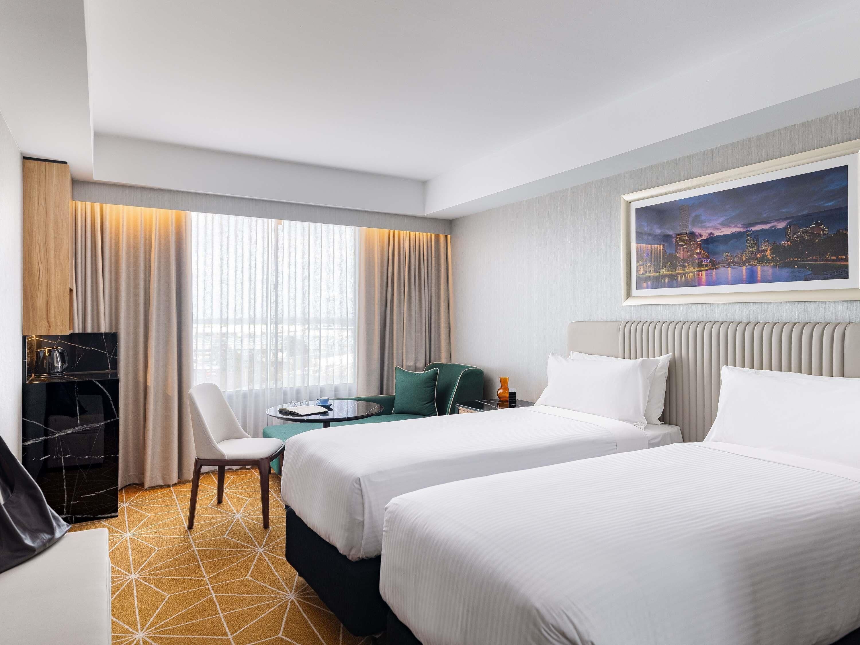 A hotel room with two beds, a desk, a chair, a coffee machine, and a cityscape painting.
