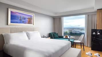 A hotel room with a large bed, a desk, a chair, a view of the city, and a painting on the wall.
