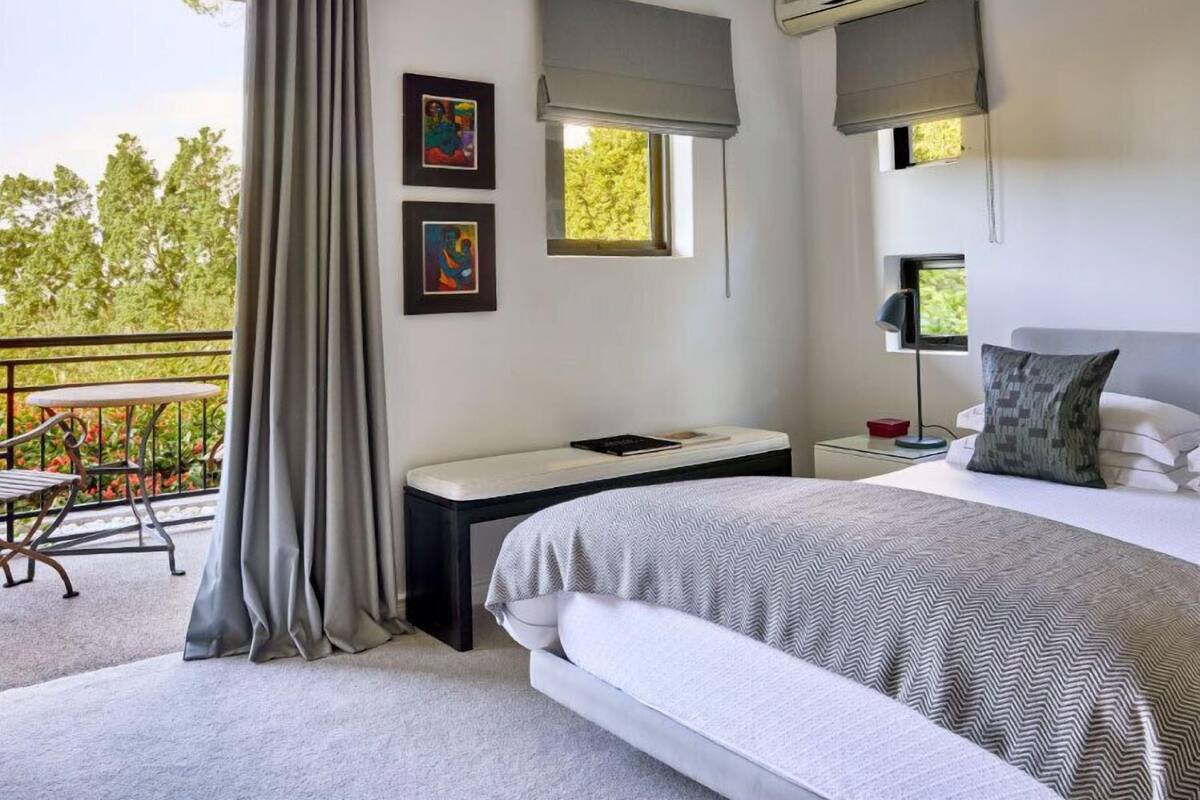 A modern bedroom with a bed, bedside table, and a desk. There are two windows with curtains, an air conditioning unit, and a balcony with a table and chairs.