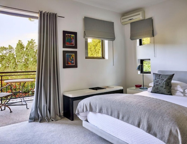 A modern bedroom with a bed, bedside table, and a desk. There are two windows with curtains, an air conditioning unit, and a balcony with a table and chairs. - Kensington Place (Cape Town)