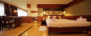 A hotel room with a large bed, wooden headboard, bedside lamps, a desk with a chair, and a dining area with a table and chairs.
