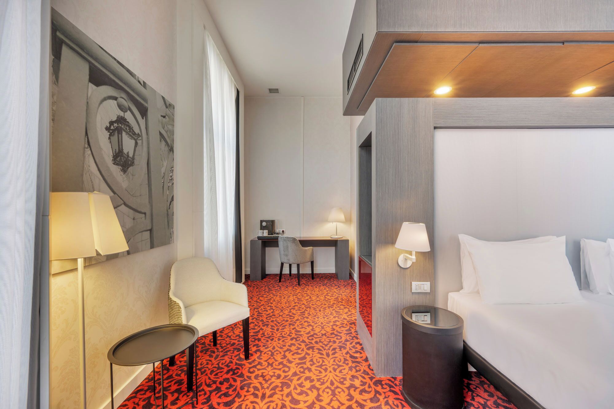 A hotel room with a bed, a desk with a chair, a lamp, and a patterned carpet.