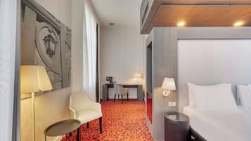 A hotel room with a bed, a desk with a chair, a lamp, and a patterned carpet.