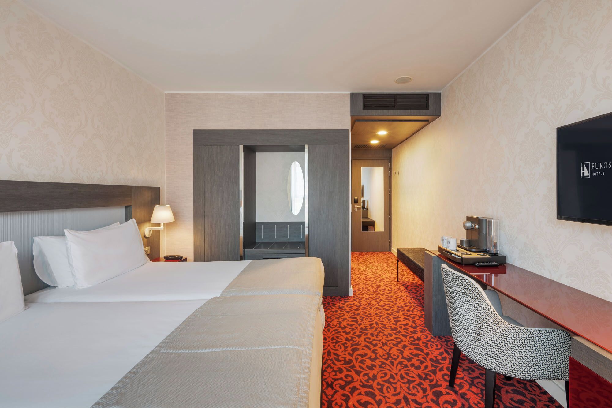 A hotel room with a bed, a desk, a chair, a TV, and a bathroom visible through an open door.