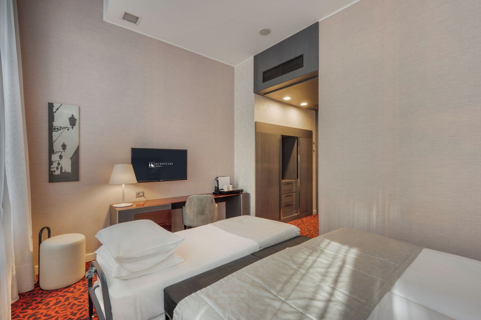 A hotel room with two beds, a desk with a lamp, a television, and a wardrobe.