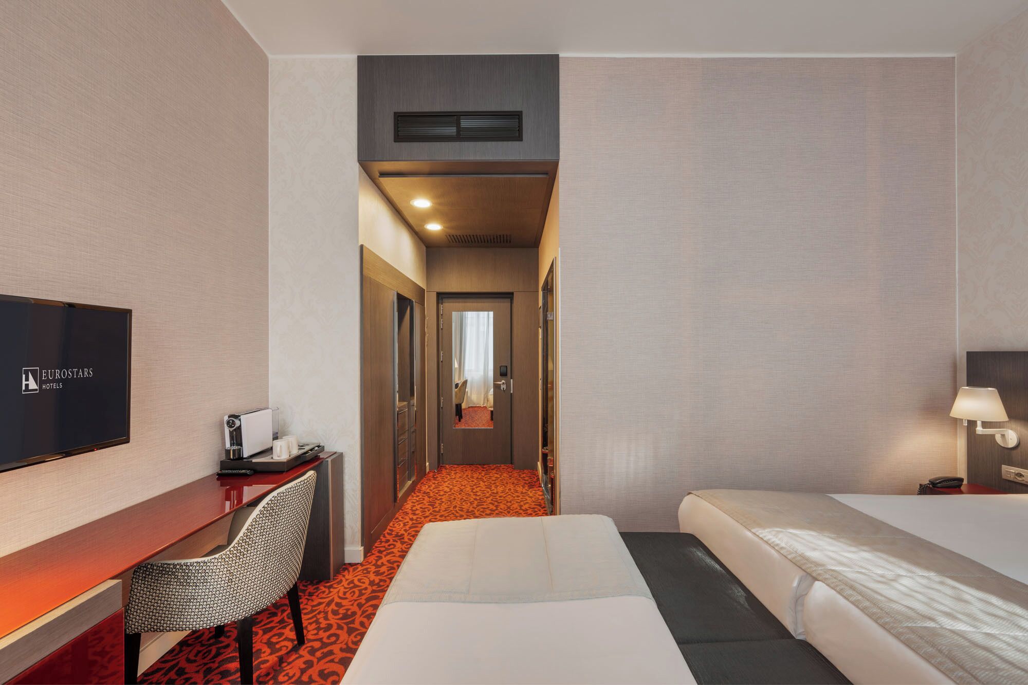 A hotel room with a bed, a desk with a chair, a television, and a bathroom visible through an open door.