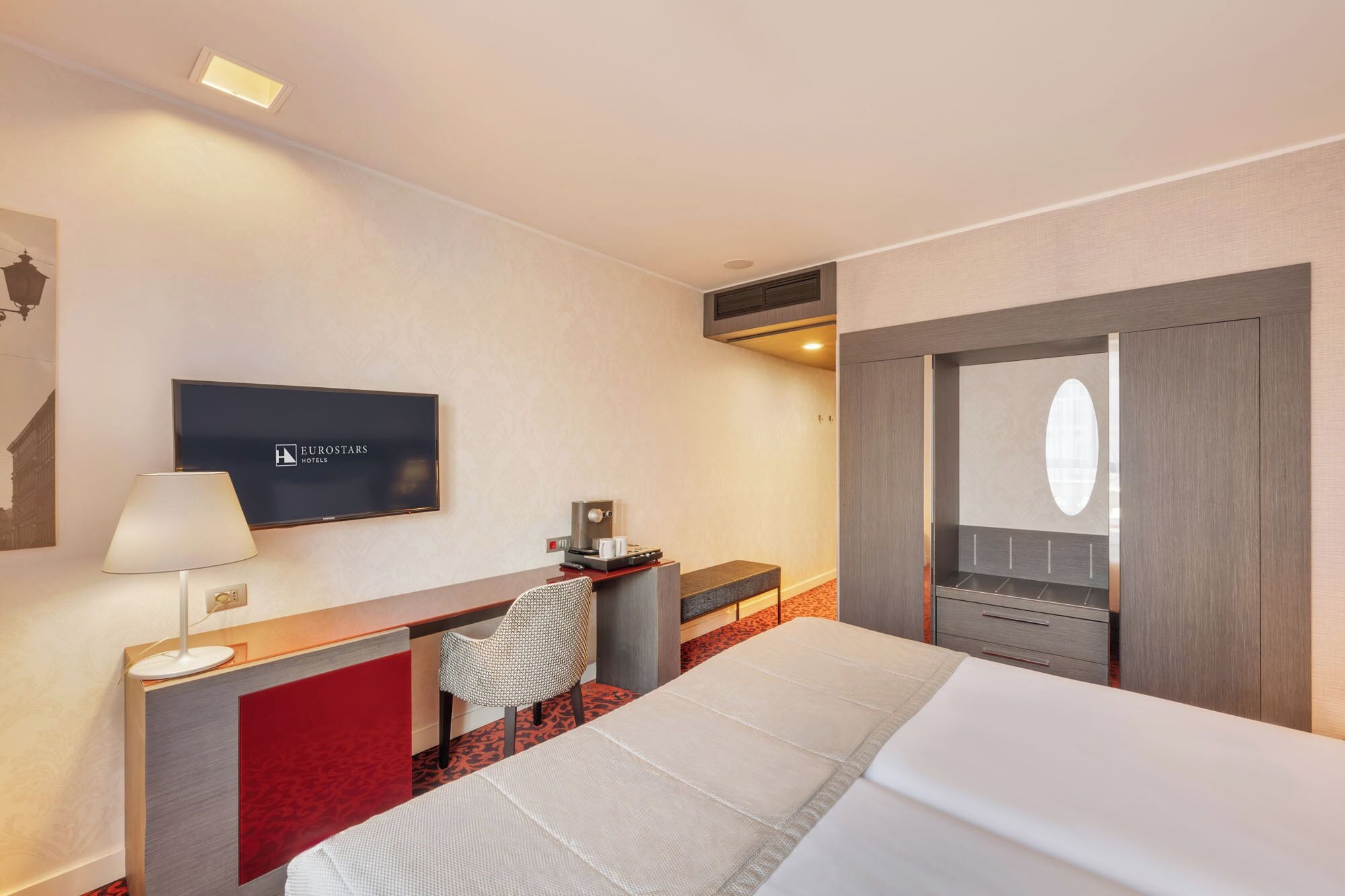 A hotel room with a bed, a desk with a chair, a television, and a wardrobe.