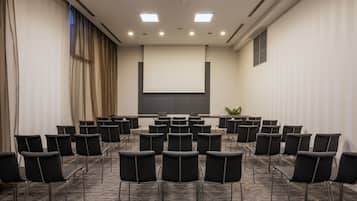 Un salón de conferencias con filas de sillas negras, una pantalla grande y suelo alfombrado.