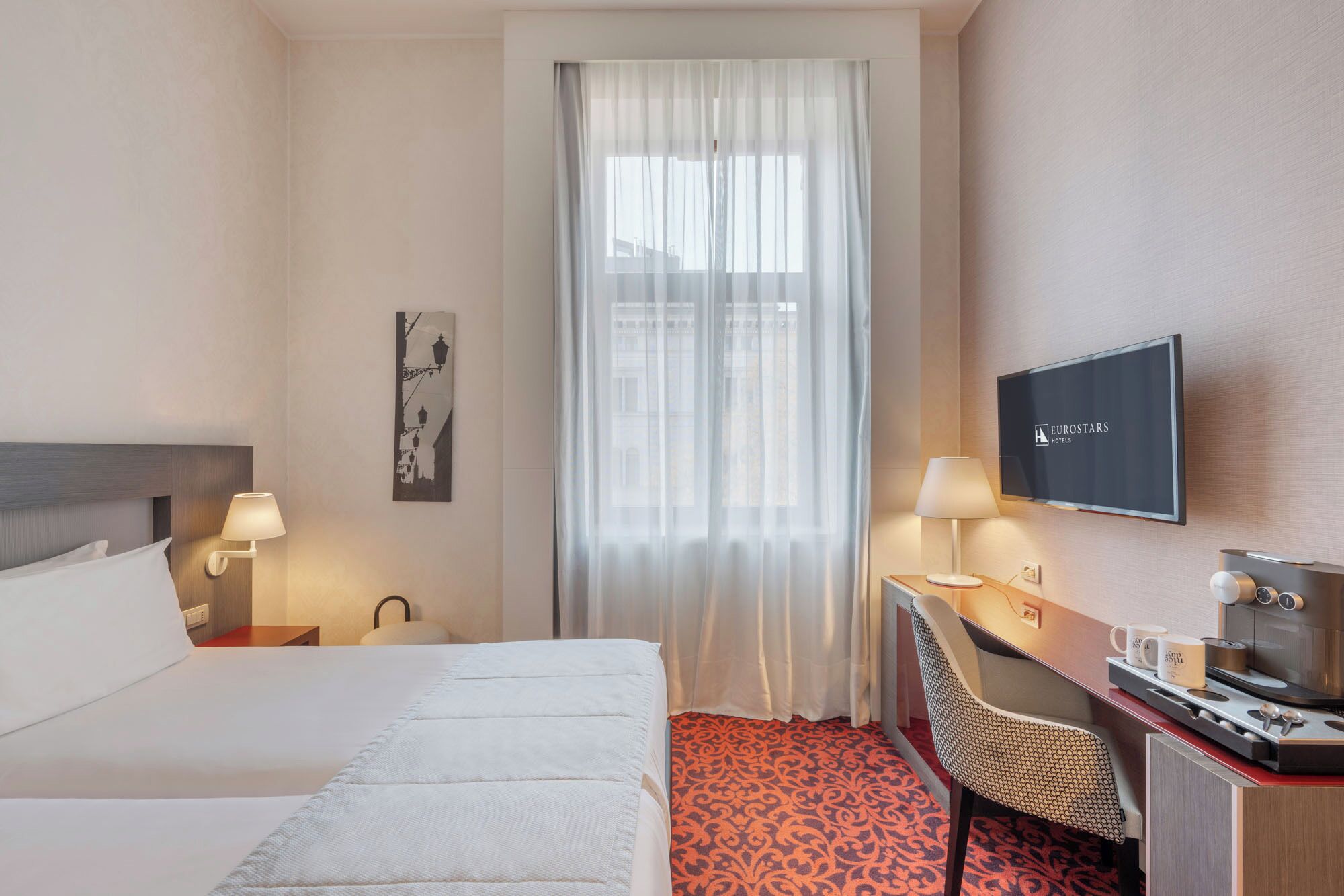 A hotel room with a bed, a desk with a chair, a television, and a window with curtains.