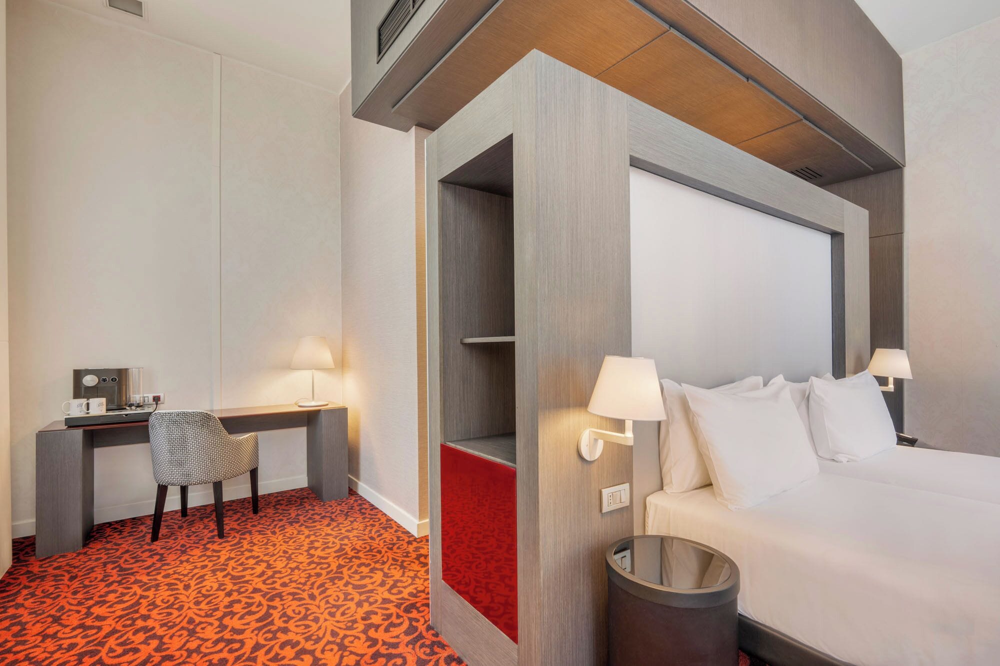 Habitación de hotel con cama, lámparas de noche, escritorio con silla, cafetera y basurero.