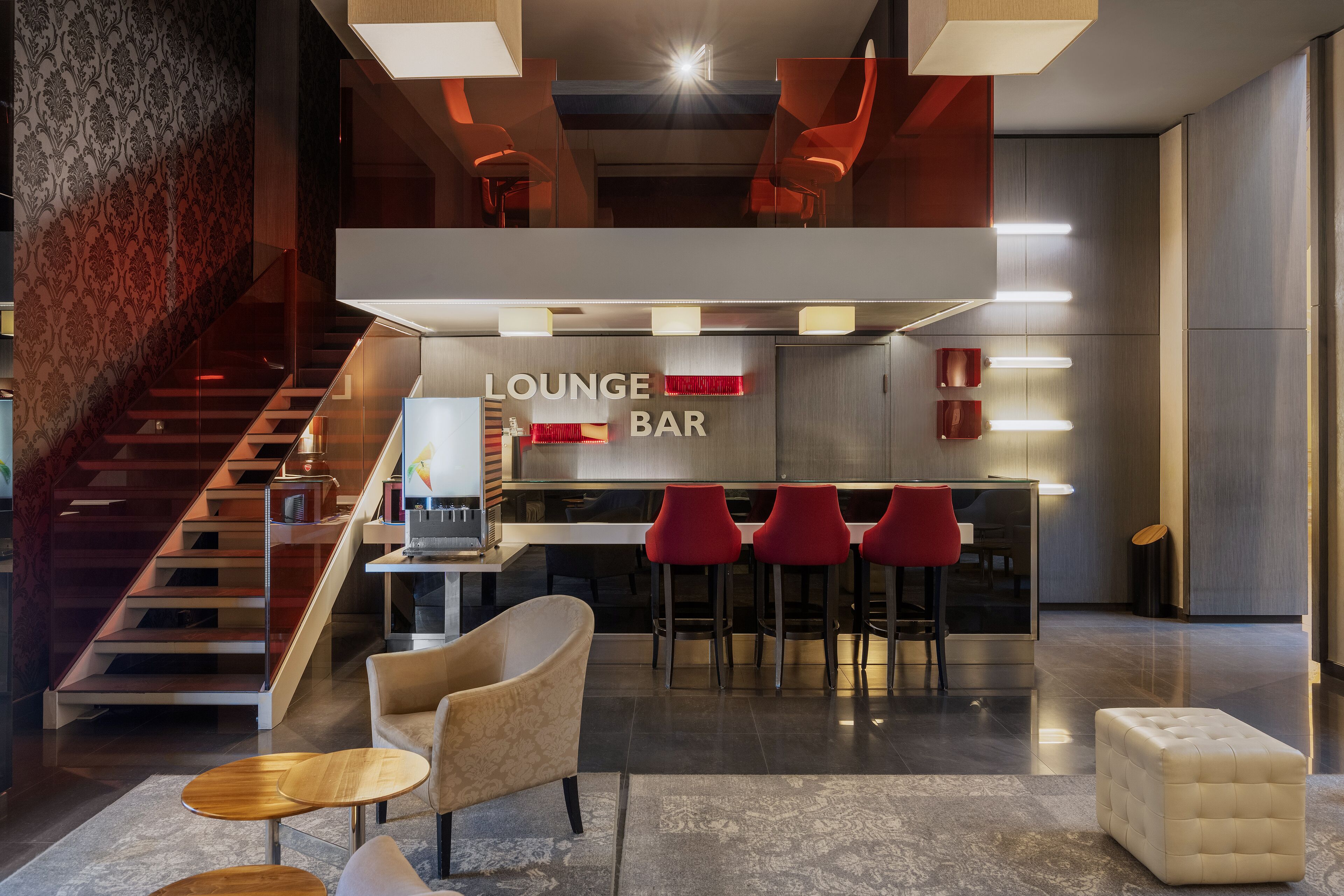 A modern lounge bar with a staircase, seating area, and bar stools.