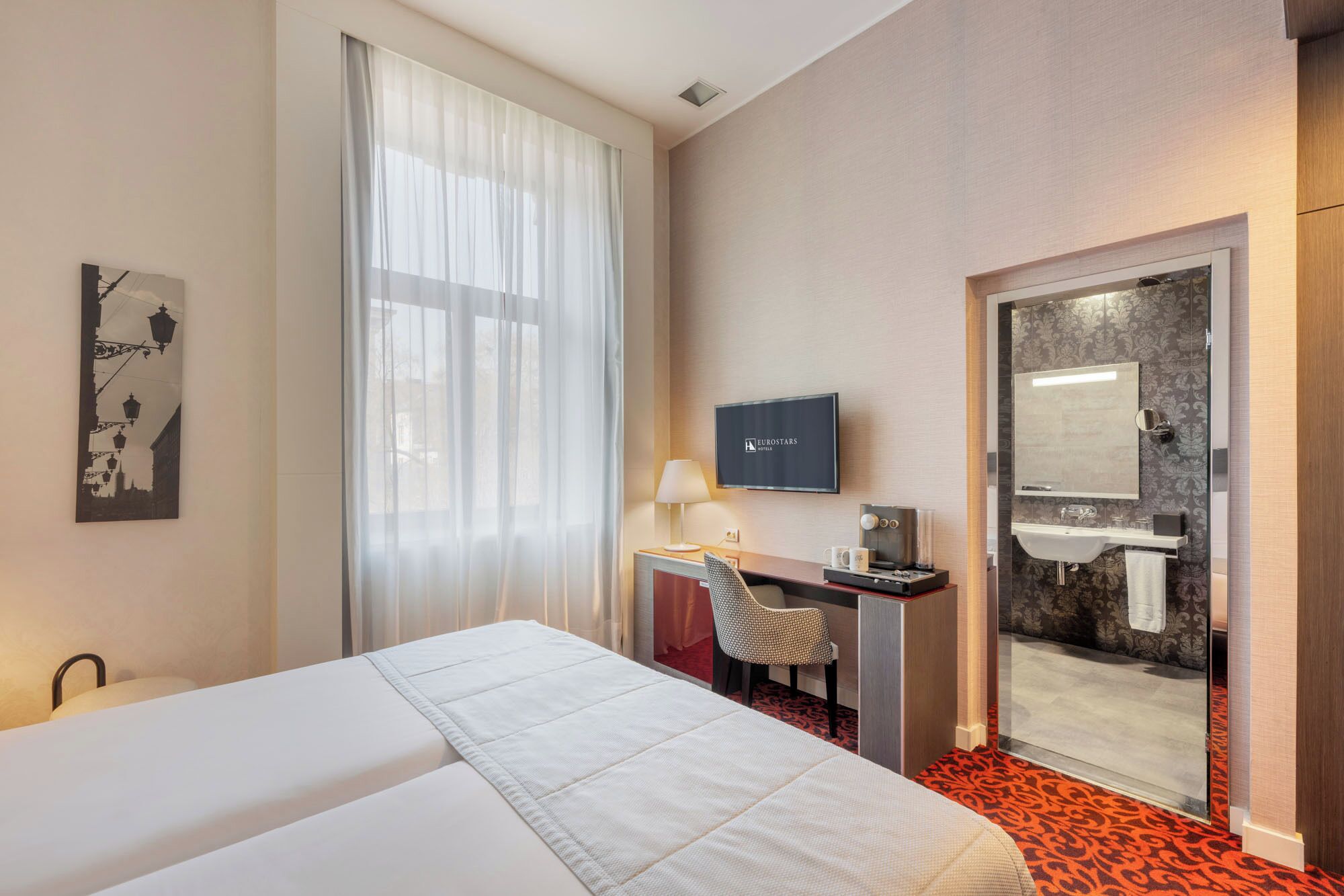 Ein Hotelzimmer mit Bett, Schreibtisch mit Stuhl, Fernseher und Badezimmer mit Spiegel und Waschbecken.