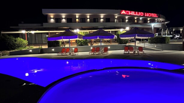 A hotel with a swimming pool, outdoor seating with umbrellas, and a well-lit facade. - Achillio Hotel (Komotini)