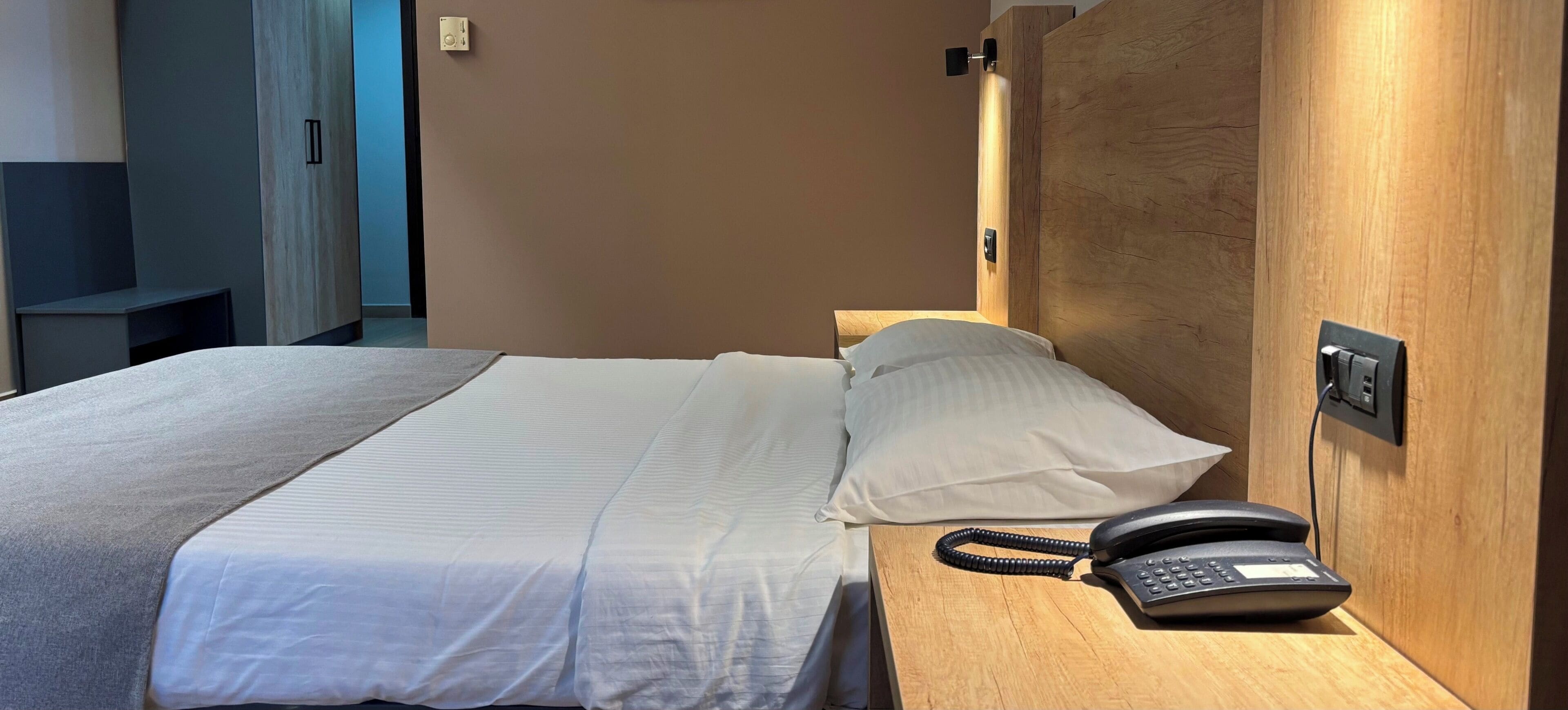 Una habitación de hotel con una cama, teléfono en la mesita de noche y cabecero de madera.
