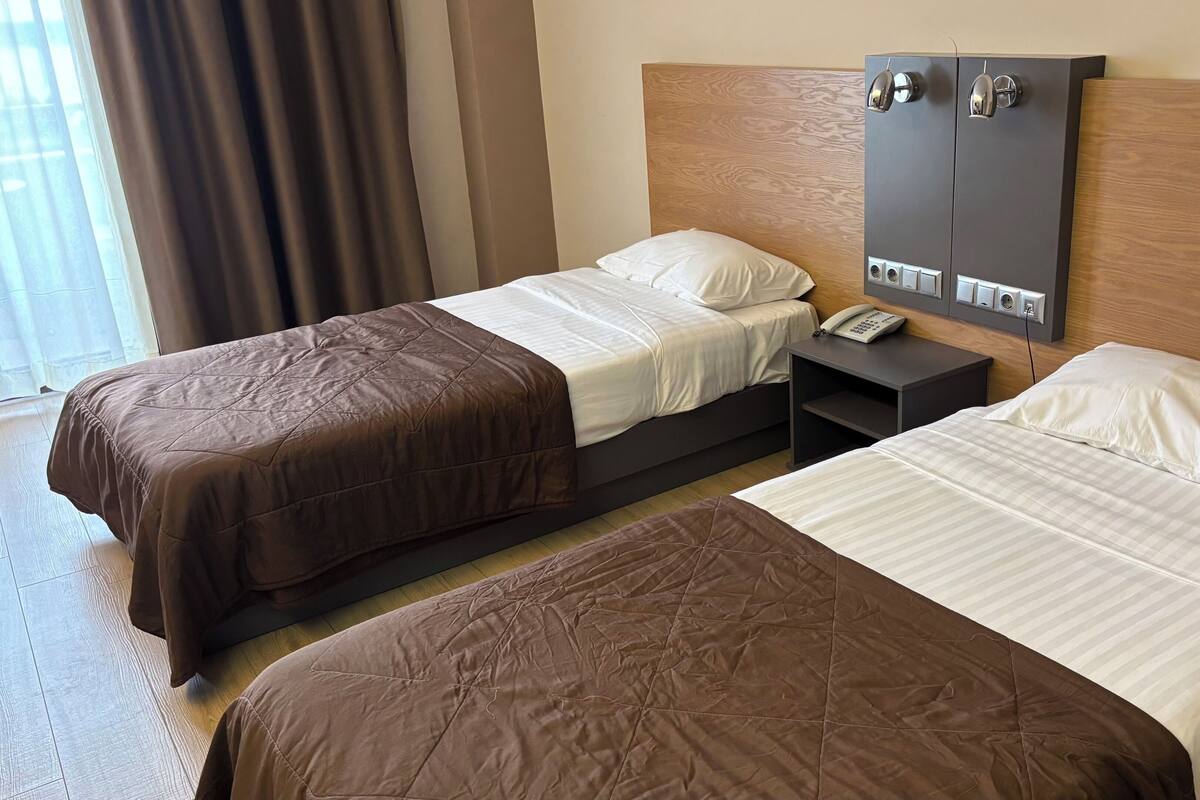Dos camas individuales con ropa de cama marrón y blanca, un cabecero de madera, una mesita de noche con un teléfono y un panel eléctrico en la pared.