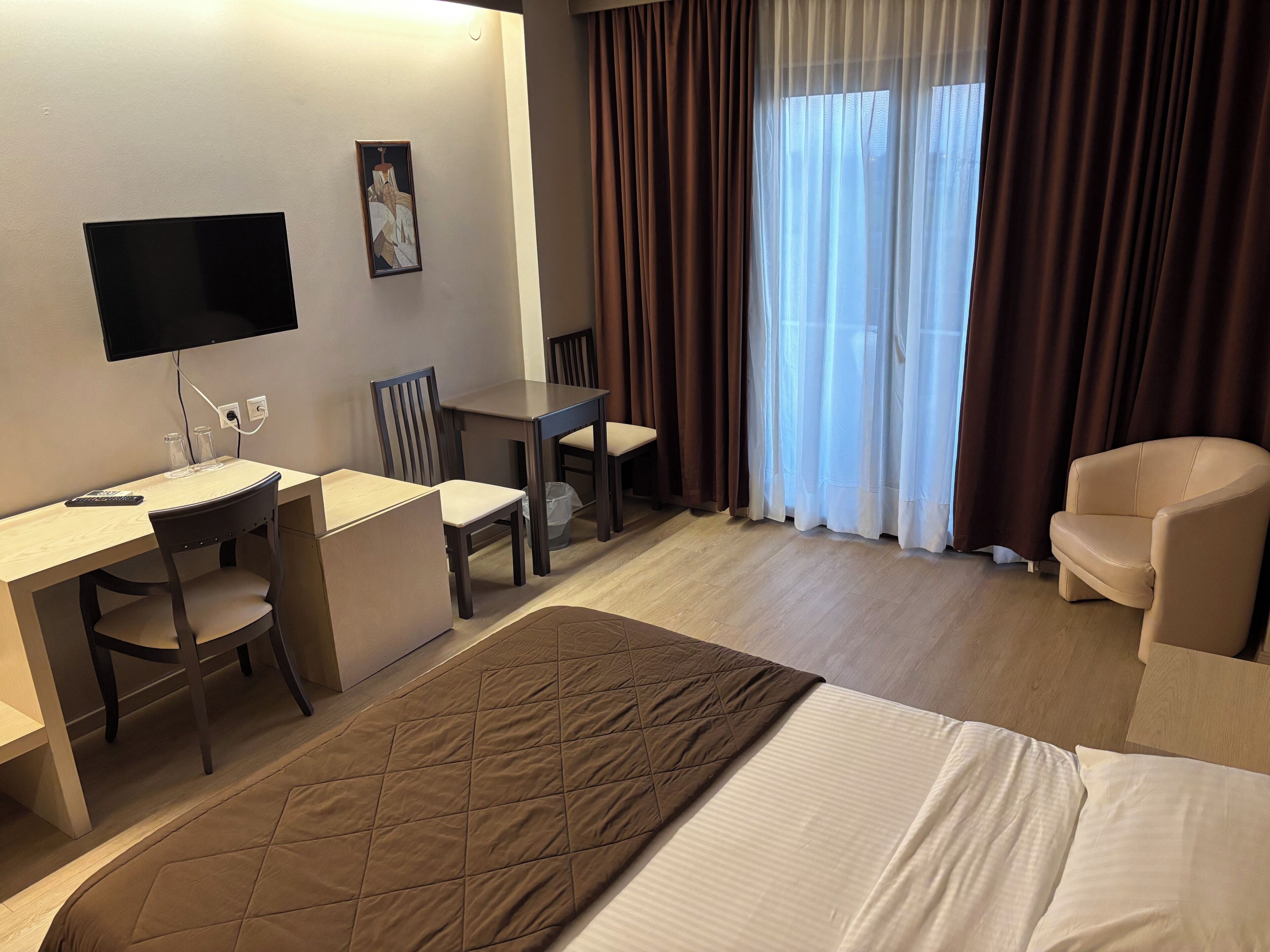 A hotel room with a bed, a desk, two chairs, a television, and a window with curtains.