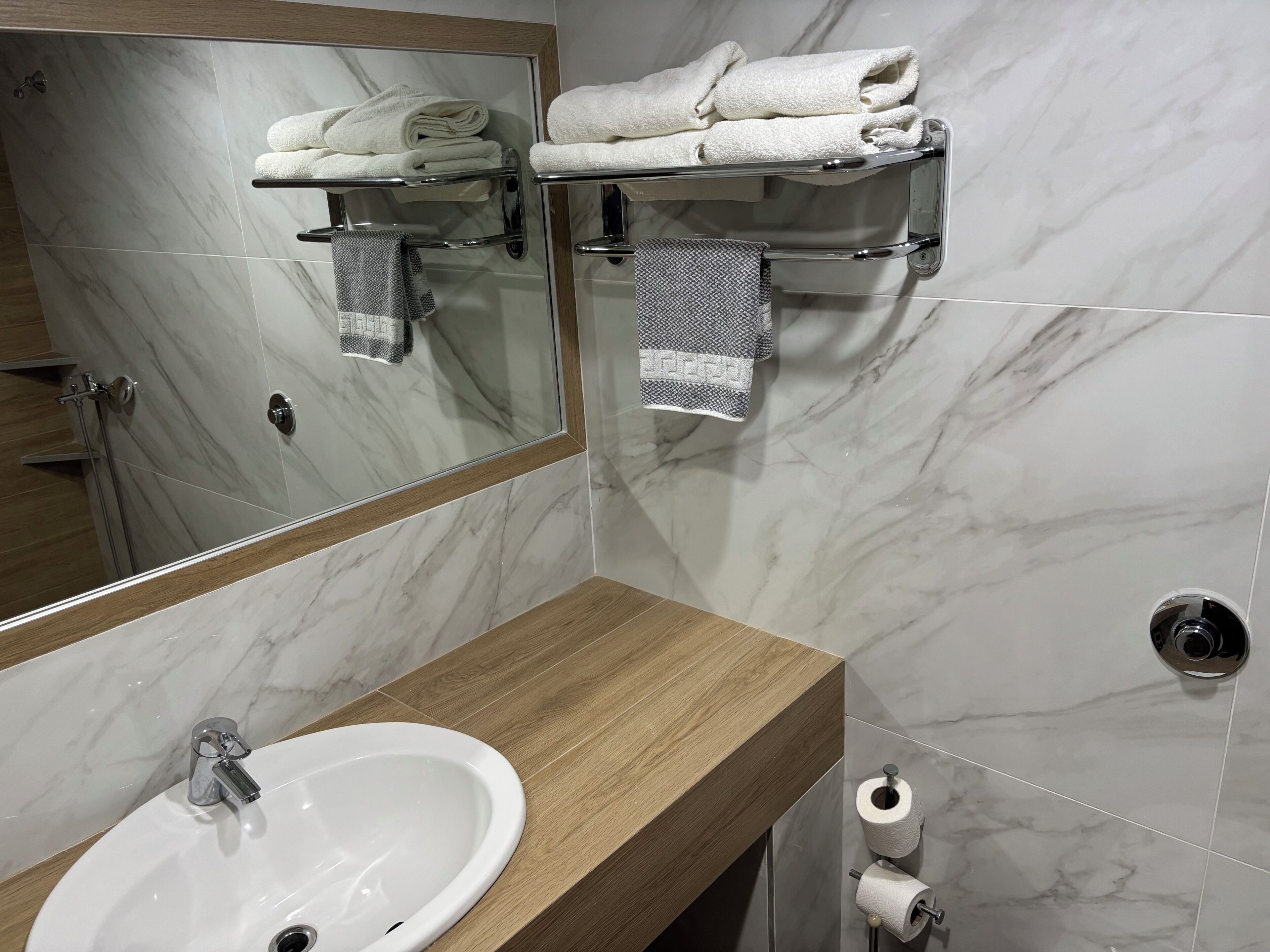 A bathroom with a marble wall, a wooden vanity, a sink, a mirror, towel racks, and a toilet paper holder.