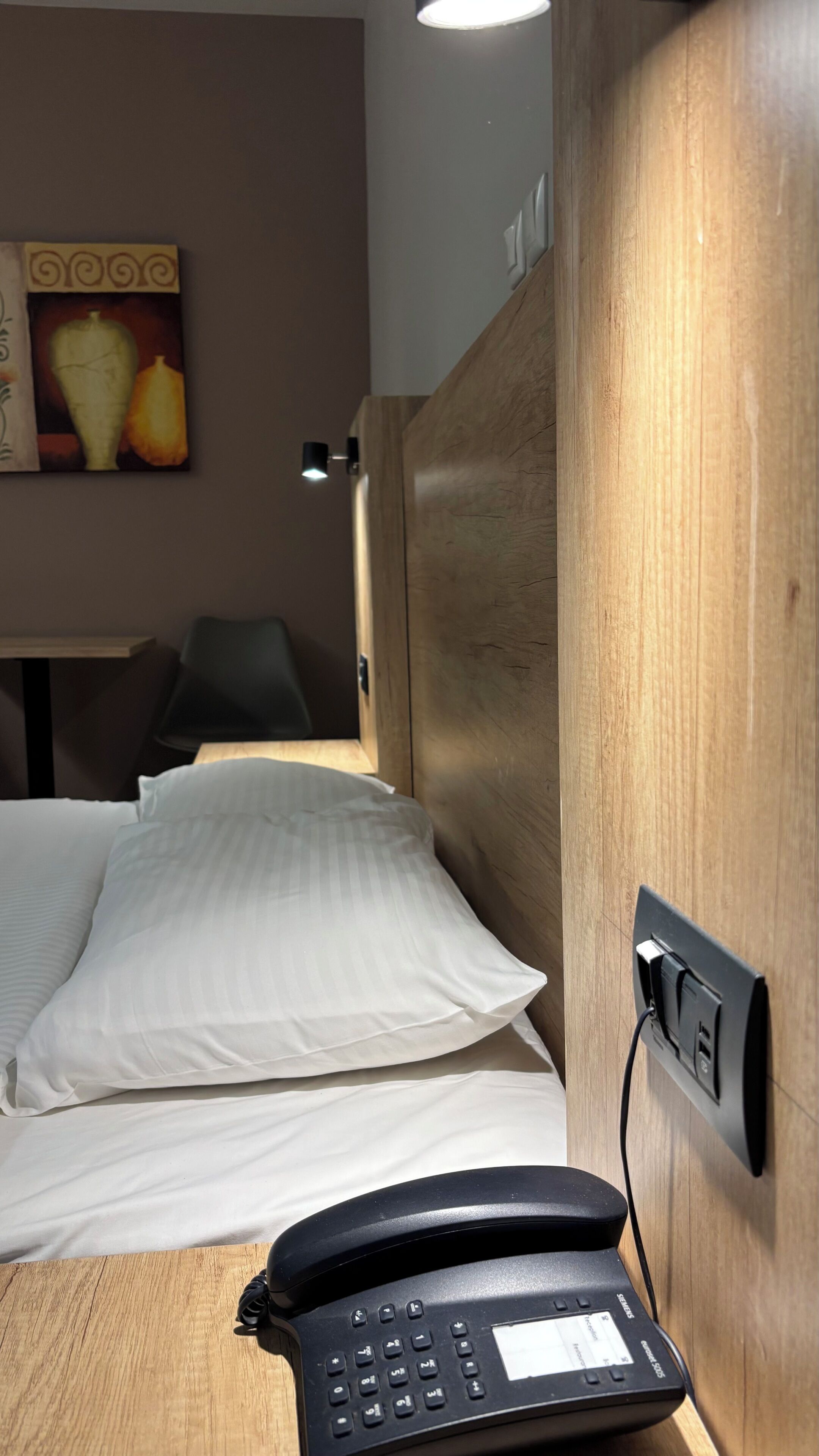 A hotel room with a wooden headboard, a bed with white linens, a black telephone, and a wall-mounted power outlet.