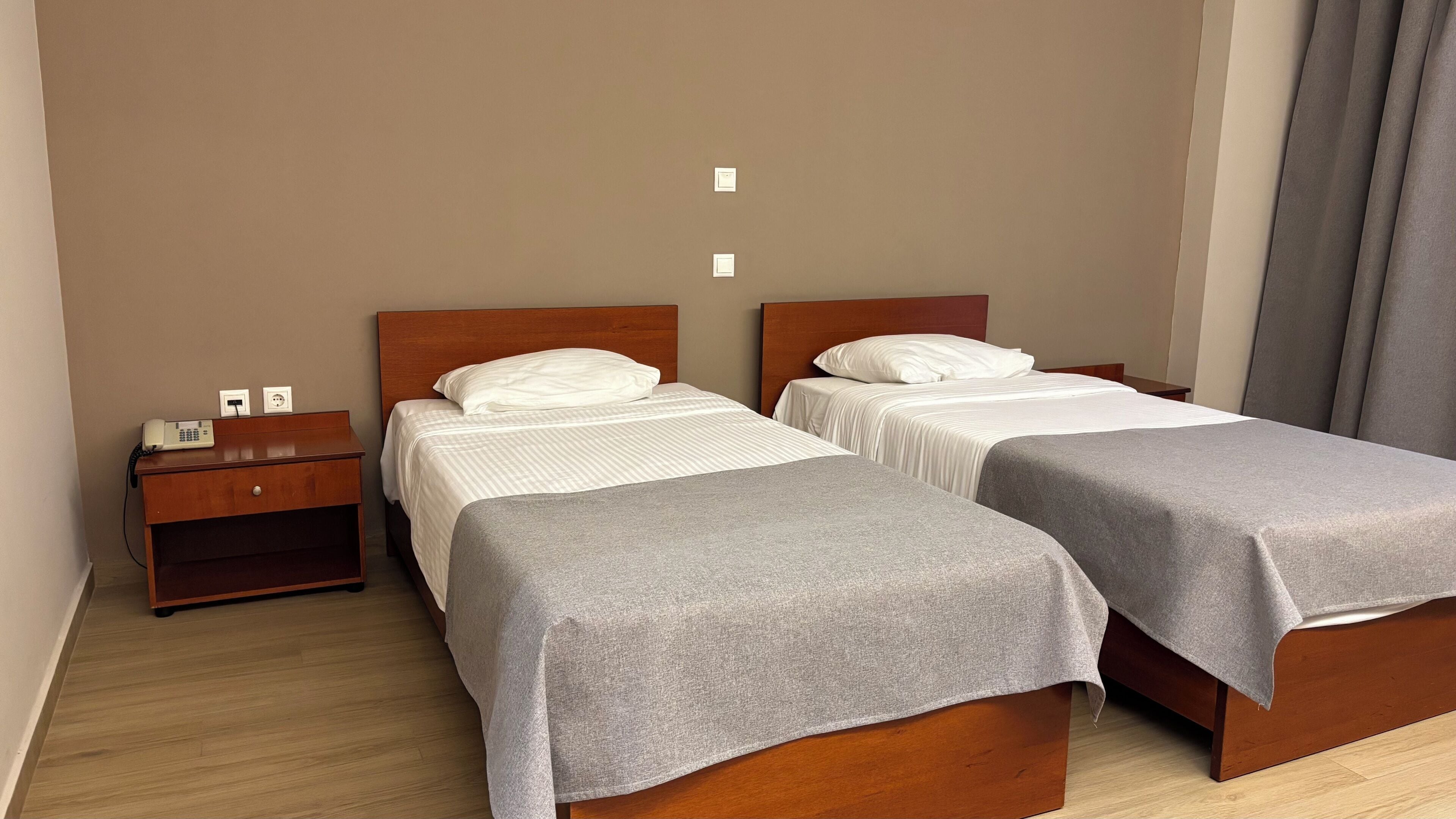 A hotel room with two single beds, a wooden nightstand with a phone, and a wall-mounted light switch.