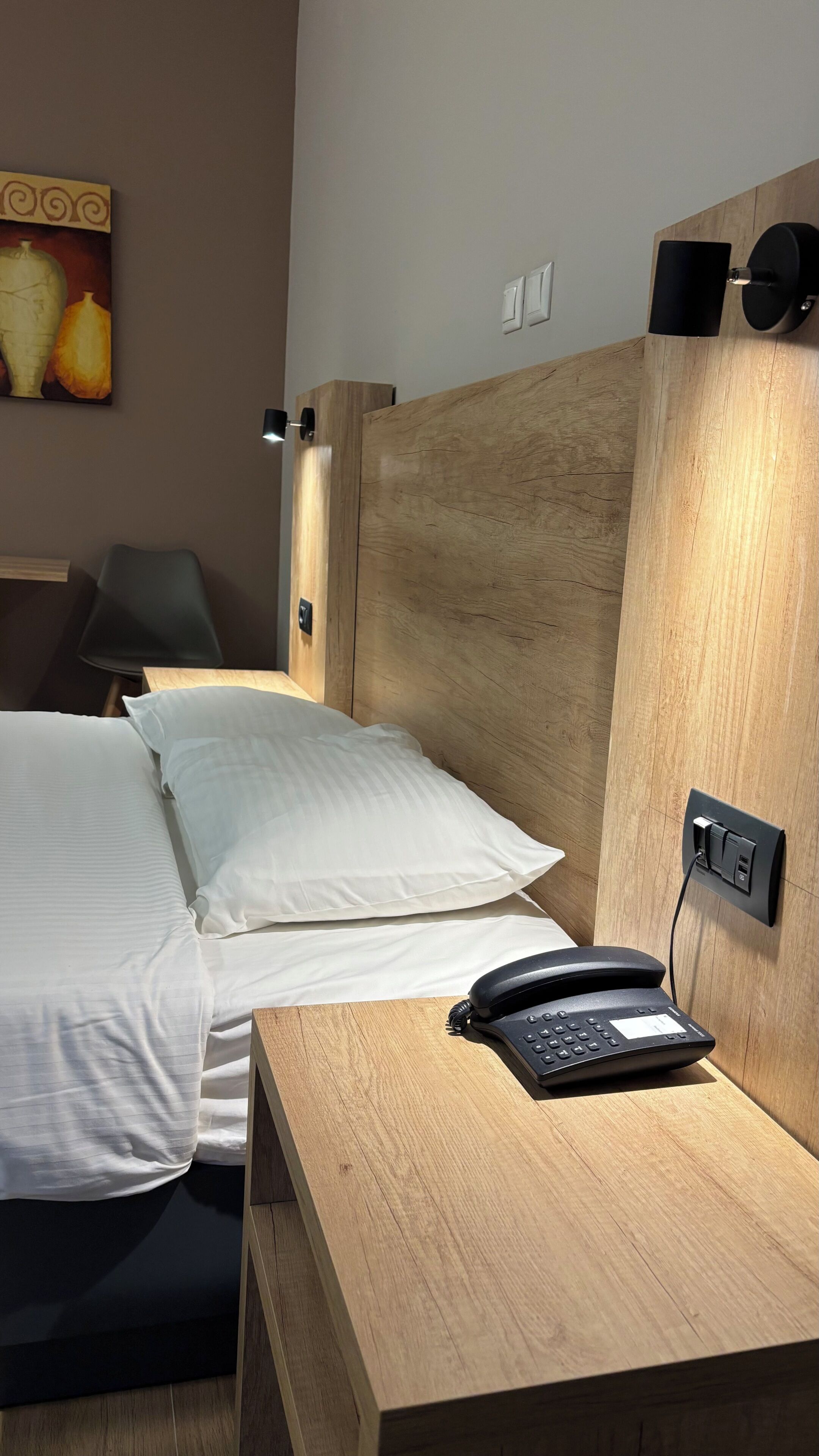 A hotel room with a wooden headboard, a bed with white linens, a bedside table with a telephone, and a wall-mounted light.