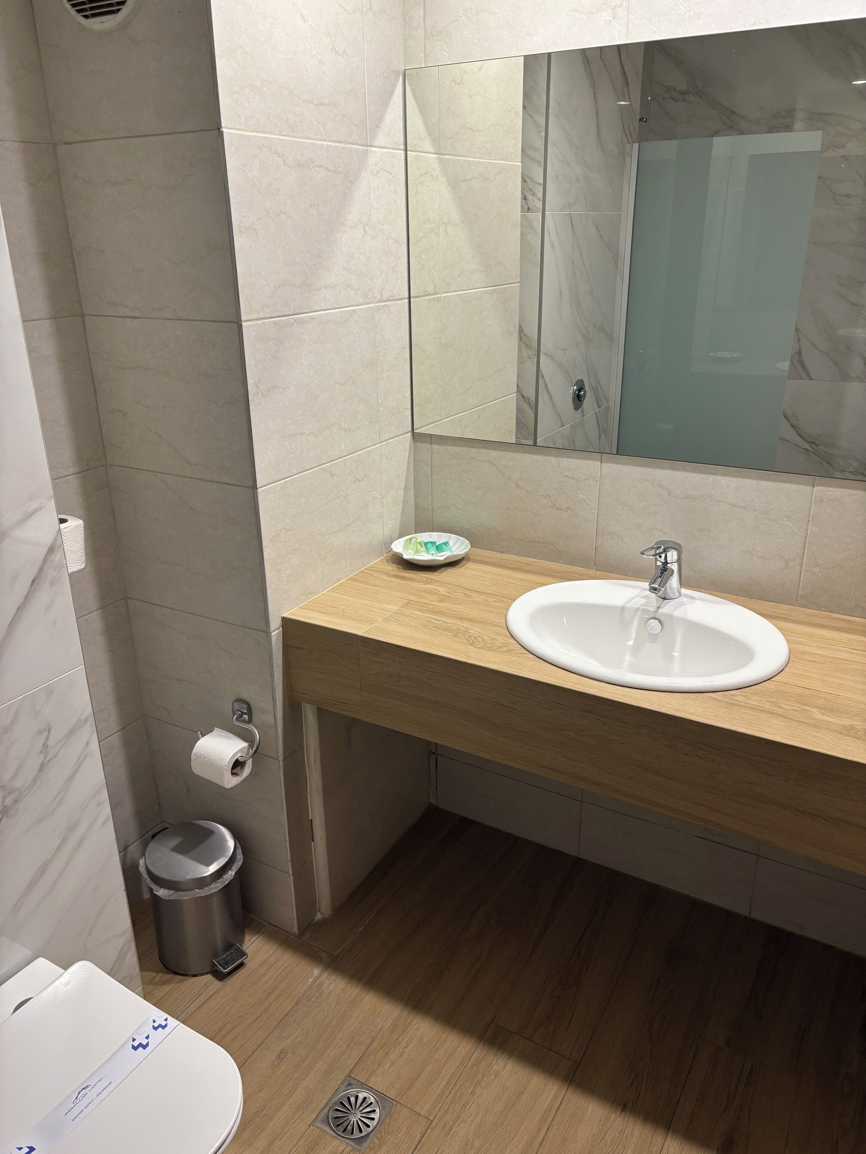Un bany amb un mur revestit de marbre, un tocador de fusta, un lavabo blanc, un mirall, un vàter i un cub de la brossa.