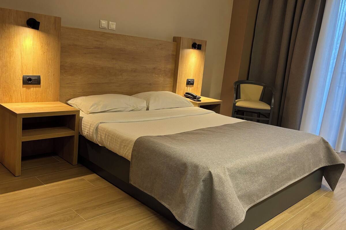 Una habitación de hotel moderna con una cama grande, cabecera de madera, mesitas de noche, una silla y vistas al exterior.