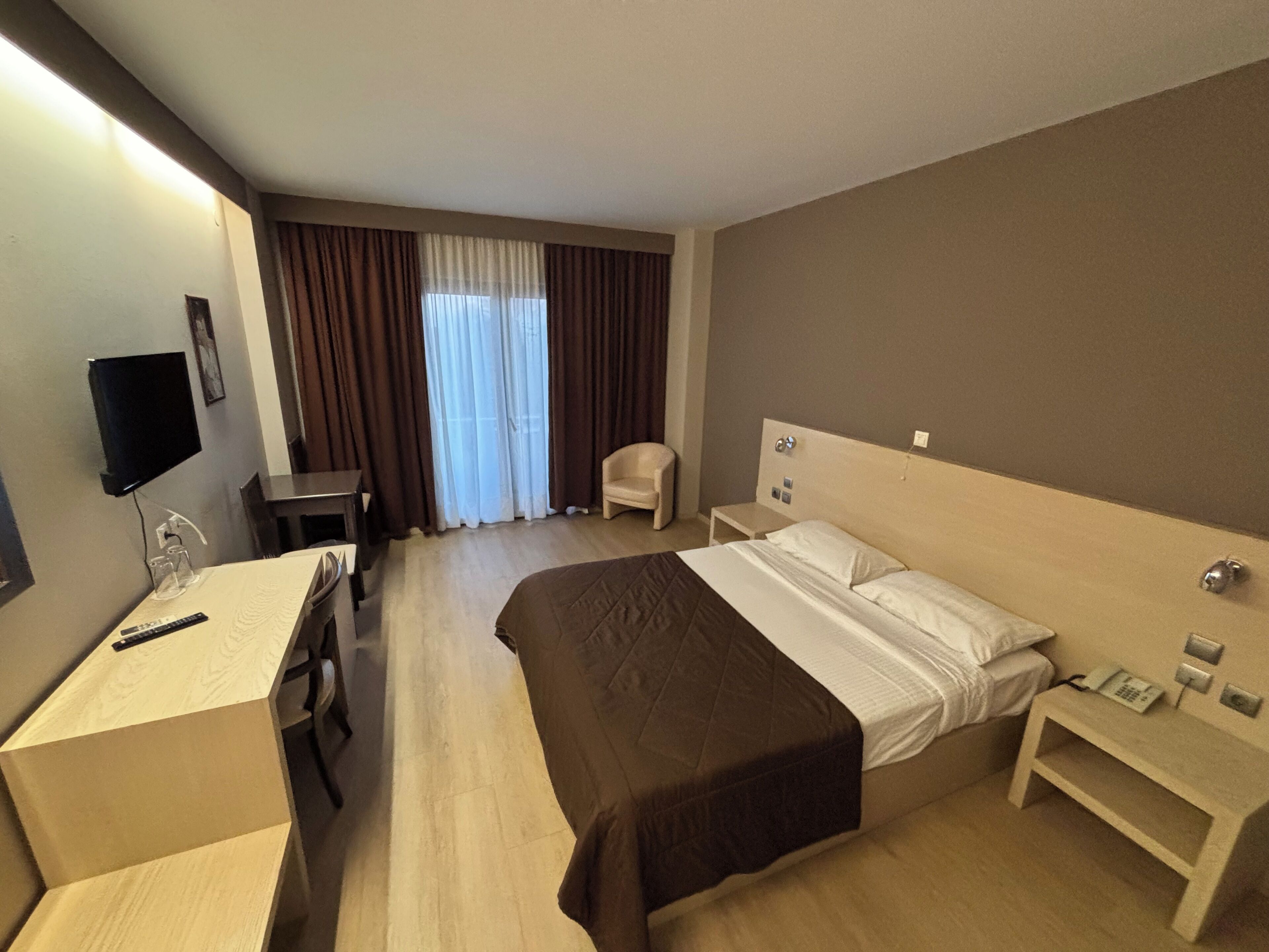 Una habitación de hotel con cama, mesita de noche, silla, escritorio, televisor y cortinas.